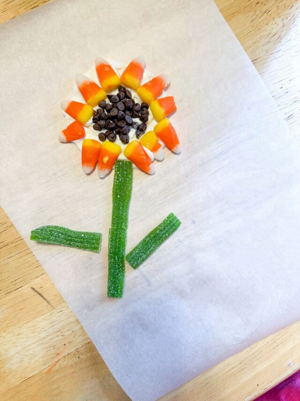 A sunflower made from candy corn and mini chocolate chips on a frosting base, perfect as an edible craft for children.